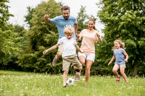 Eine Familie mit zwei Kindern spielt auf einer Wiese Fußball. Der Junge hat den Ball, alle anderen drei Personen rennen freudig hinterher.