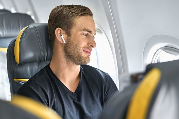 Ein etwa 30-jähriger Mann sitzt in einem Flugzeug. Er hört Musik und sieht entspannt aus dem Fenster.