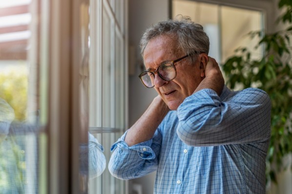 Ein etwa 70-jähriger Mann reibt sich den schmerzenden Nacken und Hinterkopf. Er steht am Fenster.