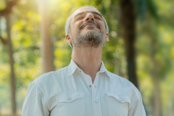 Ein etwa 45 jähriger Mann steht in der Natur und atmet tief ein. Er hat seine Augen geschlossen.