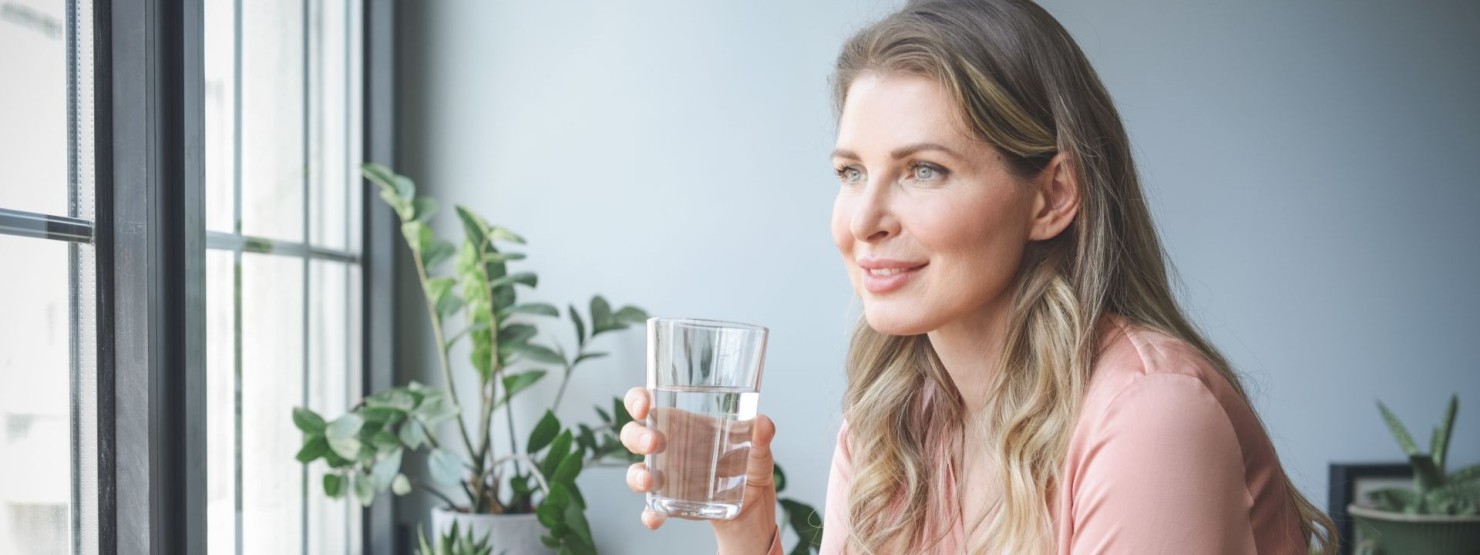 Eine etwa 45-jährige Frau hält ein Glas Wasser in der Hand und schaut lächelnd aus dem Fenster.