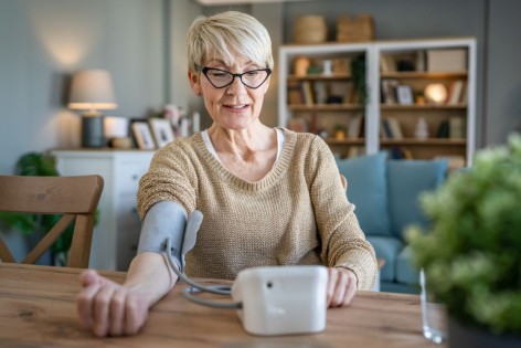 Eine etwa 60-jährige Frau misst sich mit einem Blutdruckmessgerät den Blutdruck am Arm.
