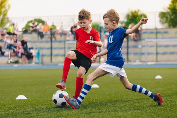 Zwei etwa 10 jährigen Jungen spielen um den Ball in einem Fussballsoiel. Beide gehören unterschiedlichen Vereinen an und tragen unterschiedliche Trikots.