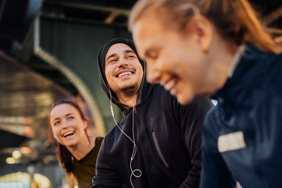 Ein junger Mann und zwei junge Frauen, alle Anfang 20, bereiten sich zusammen aufs gemeinsam Laufen vor. Sie befinden sich zum Abend in urbanem Raum udn sind guter Laune.