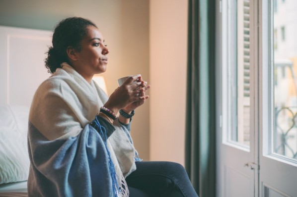 Eine etwa 40-jährige Frau sitzt mit einer Tasse auf dem Bett und schaut aus dem Fenster.