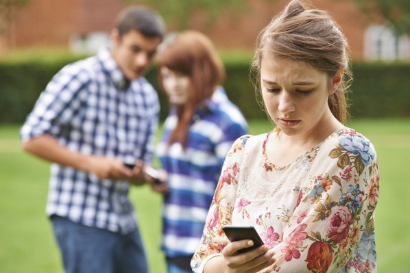 Ein etwa 15 jähriges Mädchen schaut traurig auf ihr Handy. Im Hintergrund befinden sich zwei weitere Jugendliche, die augenscheinlioch über das Mädchen im Vordergrund reden.