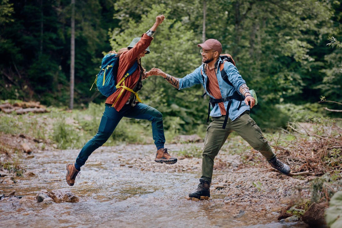 Eine Frau springt im Wald über einen Bach. Der Mann auf der anderen Seite hält helfend ihre Hand. Beide sind Mitte 20 und tragen Wanderkleidung und Rucksäcke.