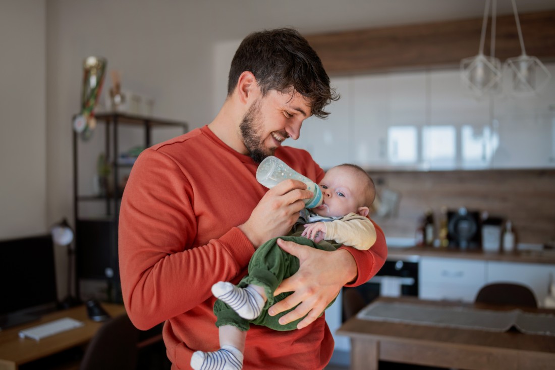 Ein etwa 30-jähriger Mann steht in seiner Wohnung. Er hält ein Baby im Arm und gibt ihm die Flasche. Er ist auf das Baby fokussiert und lächelt. Er trägt einen orangefarbenen Pullover. Im Hintergrund sieht man die Küchenzeile.