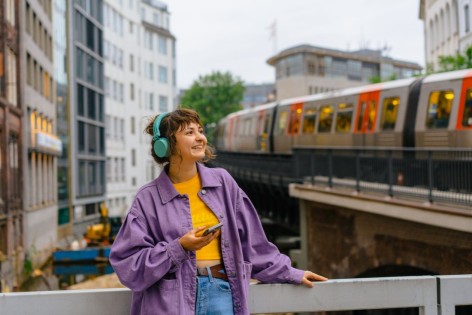 Eine etwa 20-jährige Frau hört einen Podcast über ihre Kopfhörer. Dabei steht sie mitten in der Stadt und wartet auf den Zug.