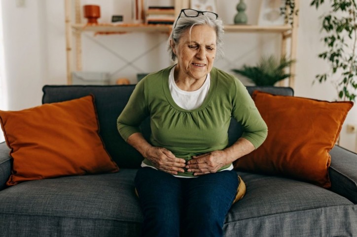 Eine etwa 60-jährige Frau sitzt auf einem Sofa und hält sich schmerzverzehrt die Hände an den Bauch.