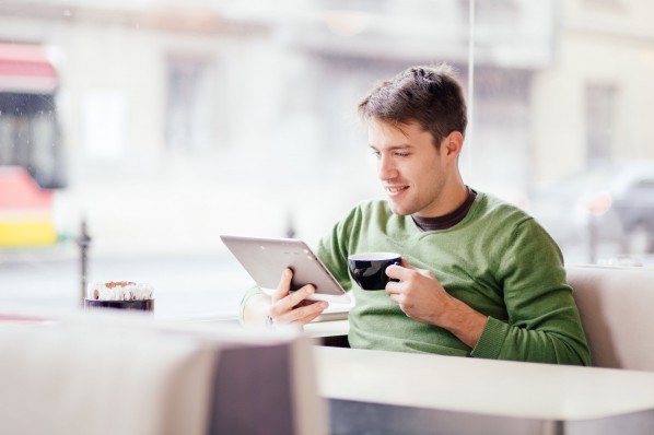 Ein etwa 30-jähriger Mann sitzt in einem Café und schaut auf ein Tablet.