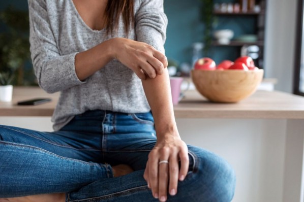 Eine etwa 30 jährige Frau sitzt auf einem Hocker in der Küche und kratzt sich am Unterarm. Die Hautstelle ist leicht gerötet. 