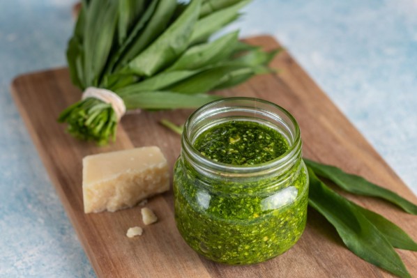 Auf einem Holzbrettchen liegt ein Bund Bärlauch, ein Stück Parmesankäse und ein Glas voll mit Bärlauch-Pesto.