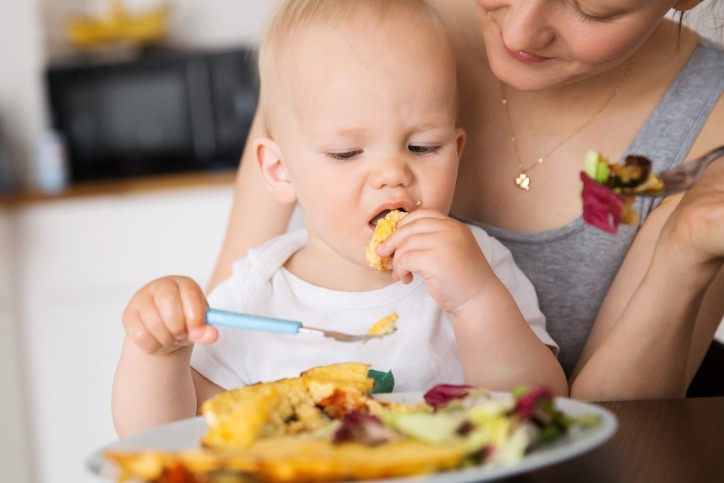 Ein Baby sitzt auf dem Schoß seiner Mutter und isst sein Essen nach der Baby Led Weaning Methode.