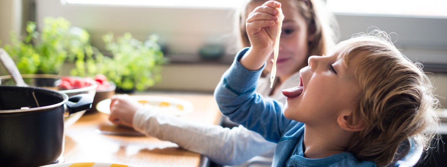 Ein etwa 4-jähriger Junge steckt sich Spaghetti in den Mund. Er sitzt dabei am Essenstisch neben seiner Schwester.