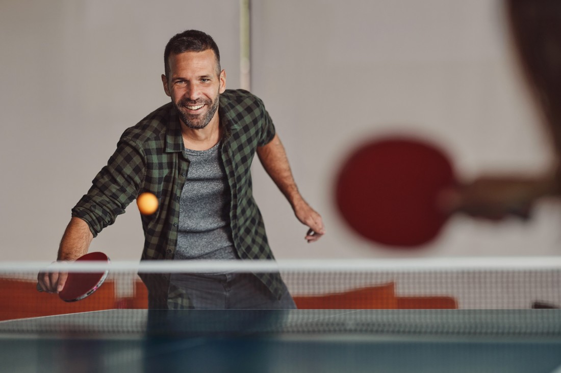 Ein etwa 45-jähriger Mann spielt in einer Halle Tischtennis. Er hat den Ball im Blick und lacht. Er trägt ein graues T-Shirt und darüber ein dunkelgrünes, kariertes Hemd mit hochgekrempelten Ärmeln.