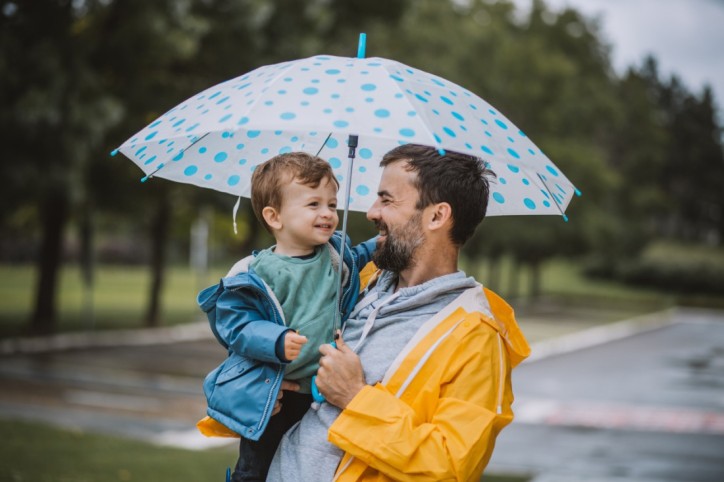 Ein Vater trägt seinen Sohn auf dem Arm und schützt ihn und sich mit einem Regenschirm