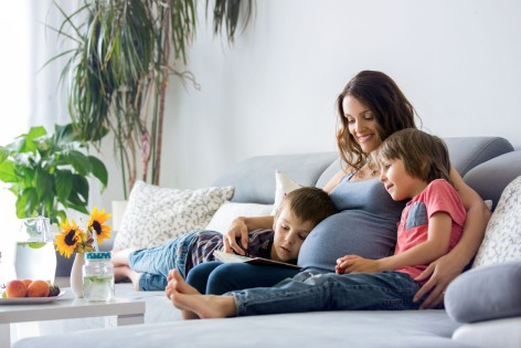 Eine schwangere Frau sitzt mit ihren zwei kleinen Söhnen auf der Couch. Alle drei schauen sich gemeinsam ein Buch an und lächeln.