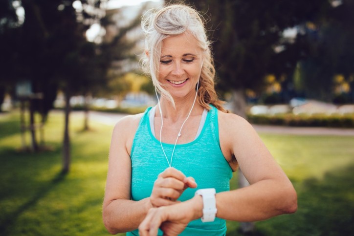 Eine etwas 55-jährige Frau macht Cardio-Training im Park und kontrolliert ihren Puls mit ihrer Fitnessuhr.