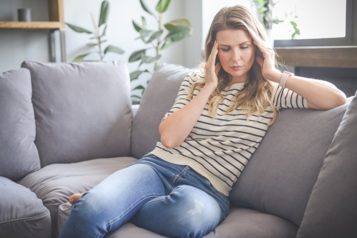 Eine Frau sitzt auf dem Sofa und leidet aufgrund von Wetterfühligkeit unter Kopfschmerzen.