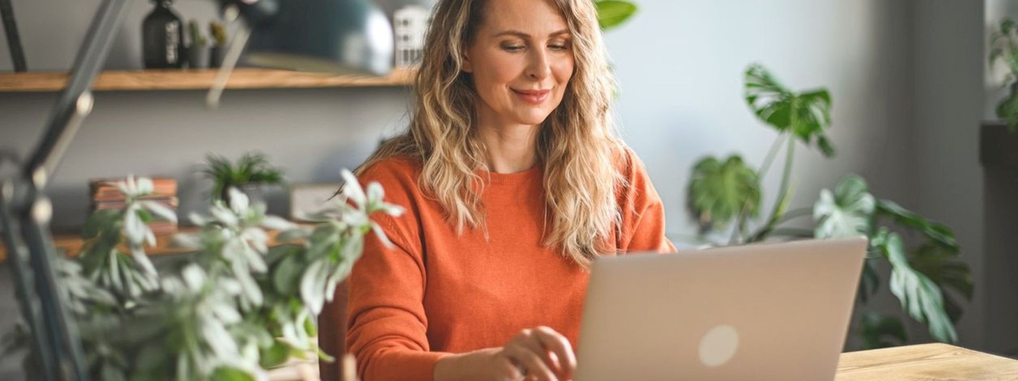 Junge Frau in orangenem Pullover sitzt in ihrem Arbeitszimmer am laptop