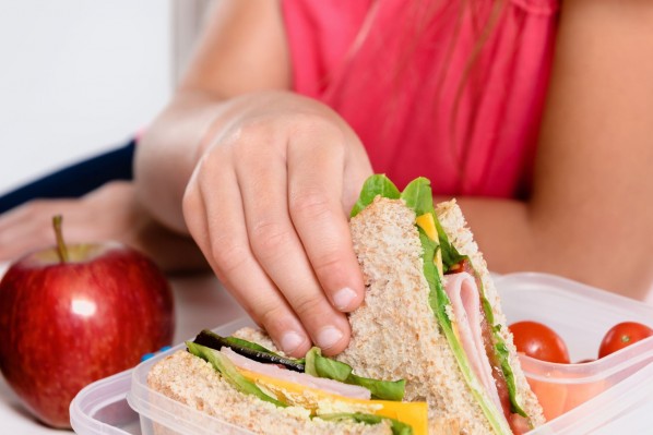 Eine Kinderhand greift nach einem belegten Sandwich in einer Tupperdose. In dieser befinden sich außerdem kleine Tomaten und daneben liegt ein Apfel.