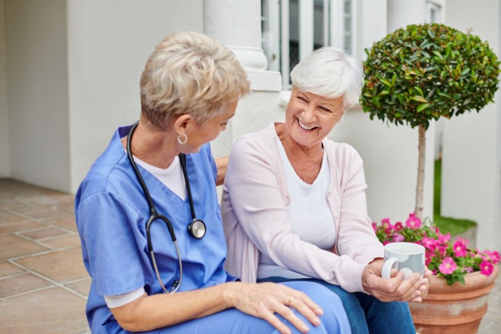 Eine Seniorin sitzt mit einer Ärztin auf einer Treppe. Die Seniorin hält eine Tasse in der Hand. Beide lächeln sich an.