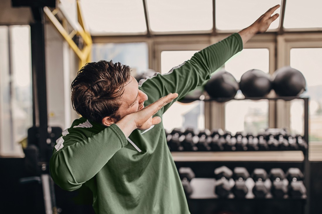 Ein energiegelandener, etwa 25 jähriger Mann steht im Fitnessstudio und macht einen Dab. Er trägt Trainingskleidung.