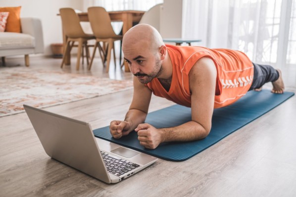 Ein etwa 45-jähriger Mann führt einen Unterarmstütz auf einer Matte Zuhause aus. Vor ihm steht ein Laptop mit weiteren Sportübungen.