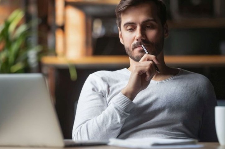 Pensive bearded man sitting at table drink coffee work at laptop thinking of problem solution, thoughtful male employee pondering considering idea looking at computer screen making decision