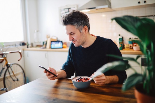 Ein etwa 40-jähriger Mann sitzt beim Frühstück und schaut auf sein Smartphone.