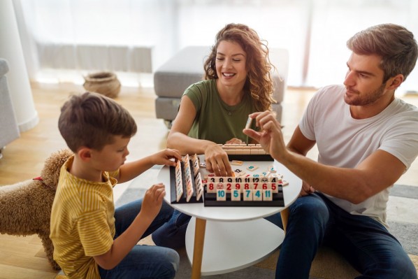 Eine dreiköpfige Familie sitzt gemeinsam an einem kleinen Tisch und spielen ein Spiel. Alle lachen ausgiebig.