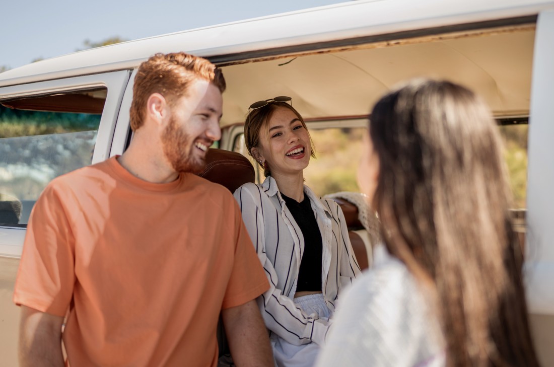 Eine junge Frau, etwa 18 Jahre, sitzt in einem alten Reisebus. Die Schiebetür des Busses ist offen. Vor dem Bus stehen ein junger Mann, etwa 20 Jahre, in einem orange-farbenen T-Shirt und eine weitere Frau mit dunklen langen Haaren, die man von hinten sieht. Die junge Frau im Bus und der junge Mann lachen. Alle sind einander zugewandt.